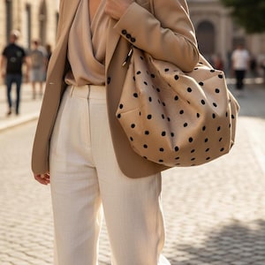 May include: A beige blazer, a light tan wrap-style blouse, white trousers, and a large beige hobo bag featuring a black polka dot pattern. The bag has a black shoulder strap and a slouchy, unstructured shape, worn over the shoulder with the blazer.