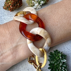 Könnte beinhalten: Ein dickes Kettenarmband mit großen, verbundenen Gliedern aus bernsteinfarbenem und weißem Harz. Das Armband hat einen goldfarbenen Verschluss.