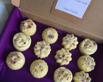 May include: A box of 12 bath melts in various shapes and sizes. The melts are white with dried flower petals on top. The box is lined with purple tissue paper. The box has a label that reads "Bath melts Cocoa and Shea butter with essential oils Just add to your bath Vegan friendly".