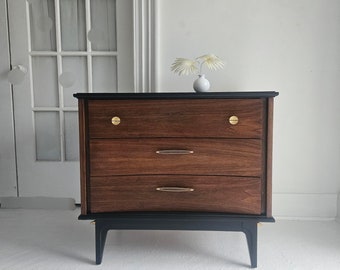 May include: A three-drawer dresser with a dark wood finish and black painted legs. The dresser has brass hardware and a simple design.