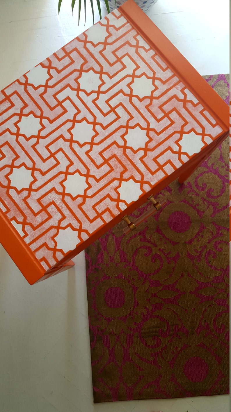 May include: Orange side table with a white geometric pattern stencil design. The table has a single drawer with a gold handle.