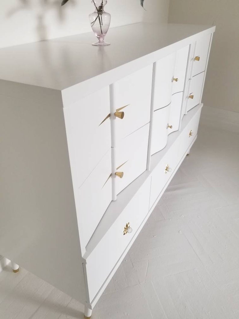 May include: White dresser with six drawers and gold-toned hardware. The dresser has a modern design with curved edges and a sleek finish.