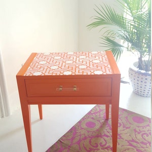 May include: Orange end table with a single drawer and white geometric pattern on the top. The table has four legs and a clear acrylic drawer pull. The table is sitting on a pink and gold patterned rug.
