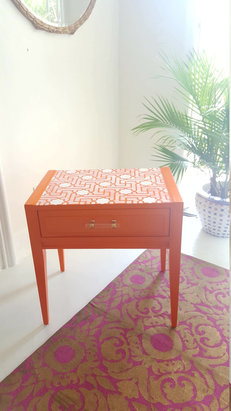 May include: Orange end table with a single drawer and white geometric pattern on the top. The table has four legs and a clear acrylic drawer pull. The table is sitting on a pink and gold patterned rug.