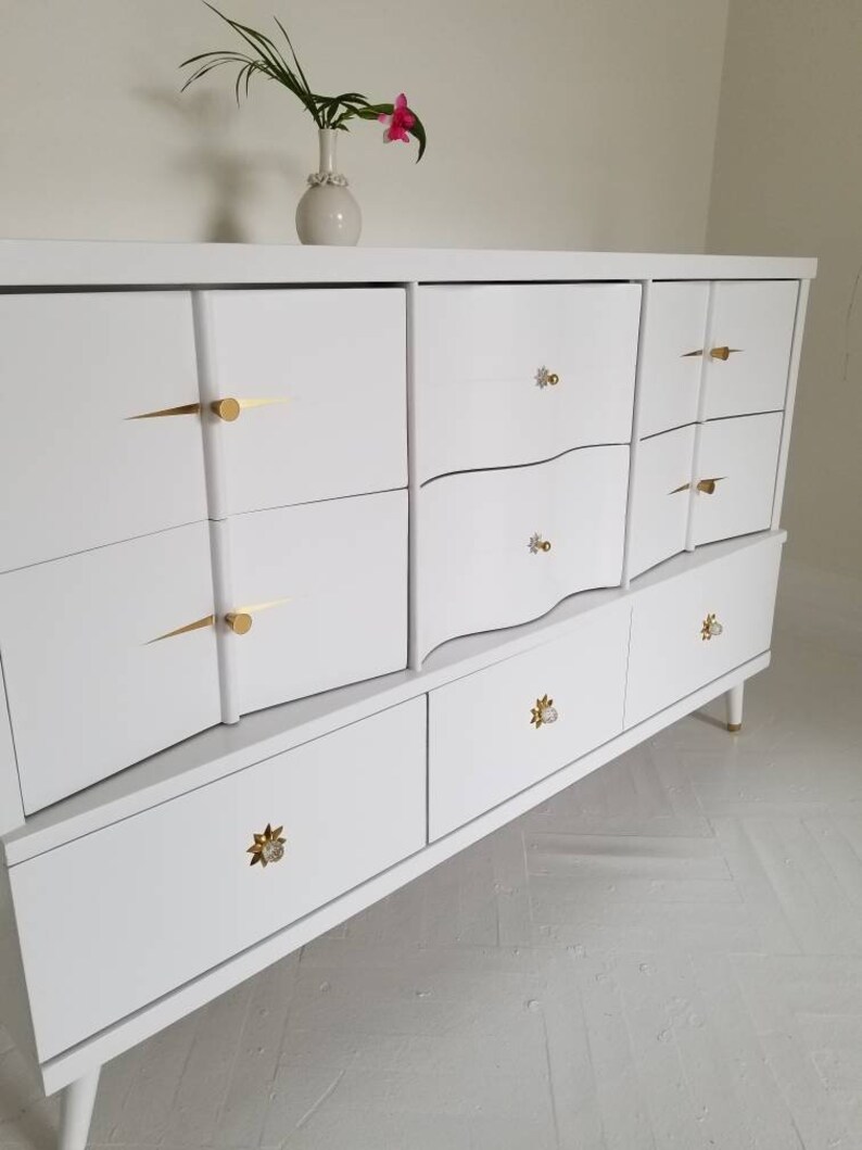 May include: White dresser with six drawers and gold-toned hardware. The dresser has a curved front and sits on tapered legs.