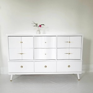 May include: White dresser with six drawers and gold-toned hardware. The dresser has a wavy design on the front and sits on four legs.