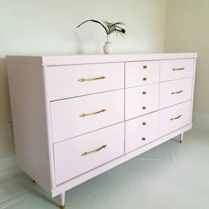 May include: A light pink chest of drawers with eight drawers and gold-toned hardware. The dresser has a modern design with tapered legs.
