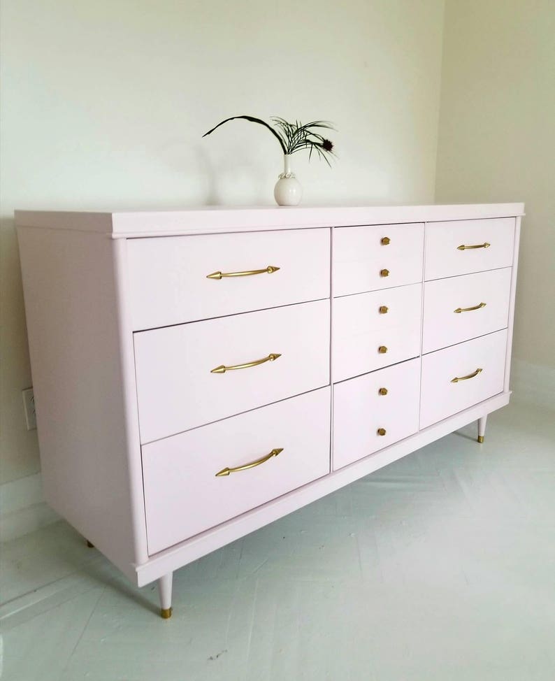May include: A light pink chest of drawers with eight drawers and gold-toned hardware. The dresser has a modern design with tapered legs.