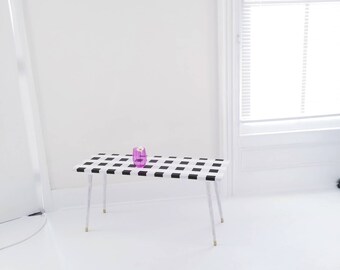May include: A white bench with black checkered top and gold legs. A purple glass vase sits on the bench.