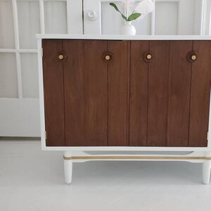 May include: A white cabinet with two wooden doors and gold accents. The cabinet has four brass knobs and sits on four white legs.