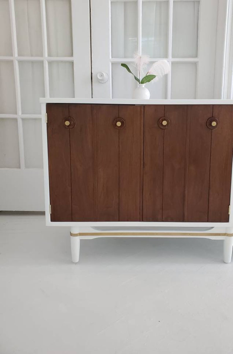 May include: A white cabinet with two wooden doors and gold accents. The cabinet has four brass knobs and sits on four white legs.