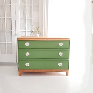 May include: A green dresser with three drawers and white ceramic knobs. The dresser has a light wood top and bottom trim.