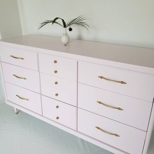May include: A light pink dresser with six drawers on each side and gold hardware. The dresser has a simple design and is made of wood.