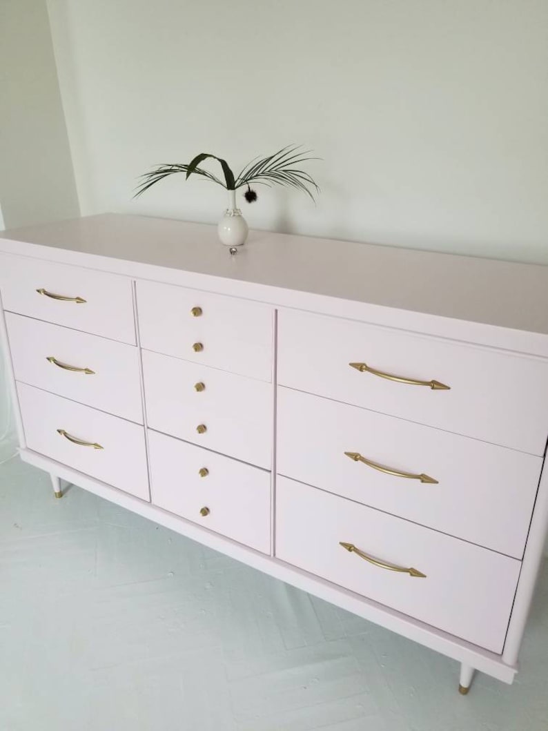 May include: A light pink dresser with six drawers on each side and gold hardware. The dresser has a simple design and is made of wood.
