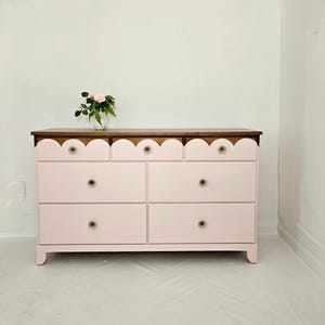 May include: A light pink dresser with seven drawers and a scalloped edge design. The top is a dark wood, and the knobs are gold. A small vase of flowers sits on top.
