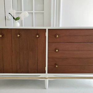 May include: A white mid-century modern dresser with four drawers and two cabinet doors. The dresser has brass hardware and a wood grain finish on the drawers and doors. The dresser is sitting on a white floor.