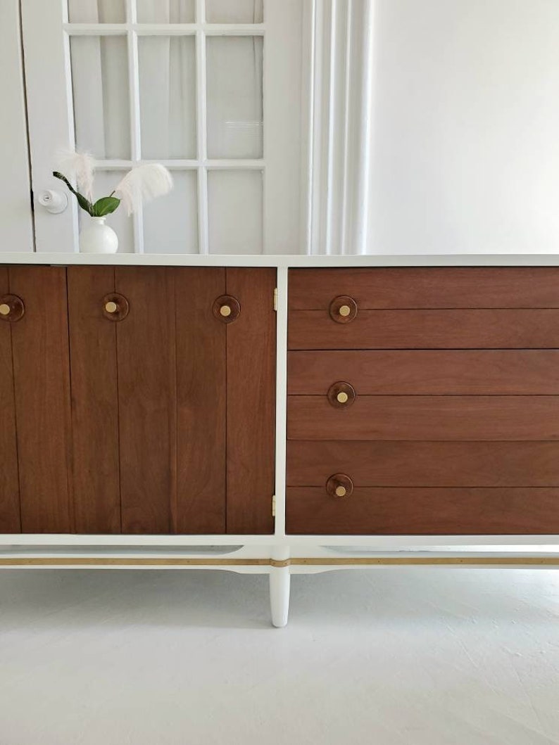 May include: A white mid-century modern dresser with four drawers and two cabinet doors. The dresser has brass hardware and a wood grain finish on the drawers and doors. The dresser is sitting on a white floor.