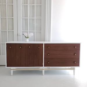 May include: A white mid-century modern dresser with brown wood drawers and gold hardware. The dresser has a white top and base with gold trim.