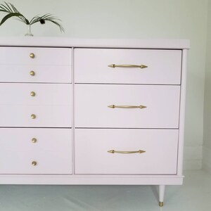 May include: A light pink chest of drawers with six drawers and gold-toned hardware. The dresser has a simple, mid-century modern design with tapered legs.