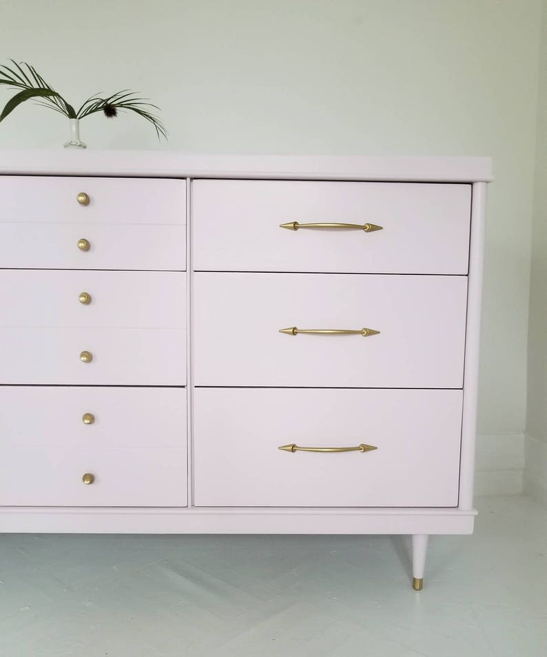 May include: A light pink chest of drawers with six drawers and gold-toned hardware. The dresser has a simple, mid-century modern design with tapered legs.