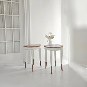 May include: Two white round side tables with brown wooden legs. The tables have a white top with a decorative pattern. A small white vase with dried flowers sits on one of the tables.