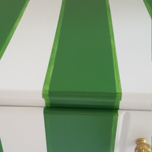 May include: A white chest of drawers with green stripes painted on the front. The dresser has three drawers with gold knobs.