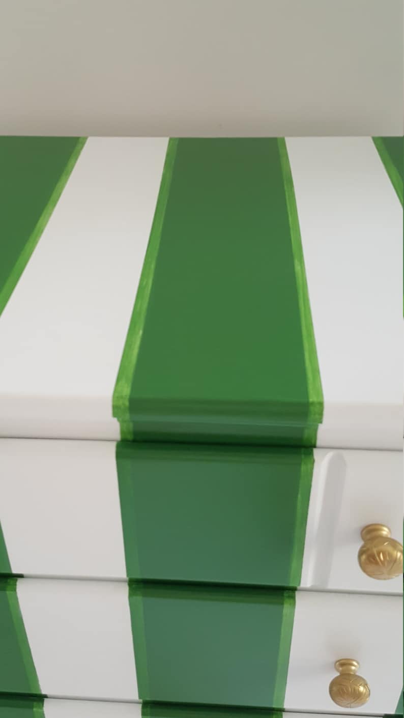 May include: A white chest of drawers with green stripes painted on the front. The dresser has three drawers with gold knobs.