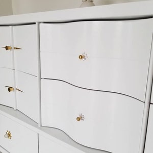 May include: White dresser with six drawers and gold and crystal drawer pulls. The dresser has a wavy design on the front.