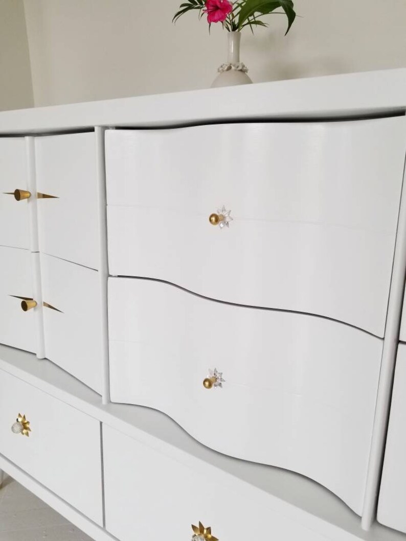 May include: White dresser with six drawers and gold and crystal drawer pulls. The dresser has a wavy design on the front.