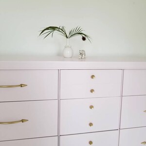 May include: A light pink chest of drawers with six drawers and gold handles. The dresser has a simple design and is made of wood. A small white vase with a green plant and a white ceramic tiger figurine are on top of the dresser.