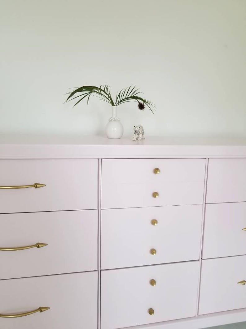 May include: A light pink chest of drawers with six drawers and gold handles. The dresser has a simple design and is made of wood. A small white vase with a green plant and a white ceramic tiger figurine are on top of the dresser.