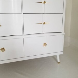 May include: White dresser with six drawers and gold star-shaped knobs. The dresser has a curved front and sits on four legs.