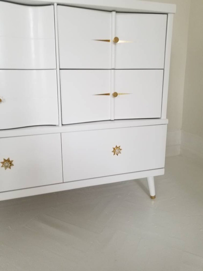 May include: White dresser with six drawers and gold star-shaped knobs. The dresser has a curved front and sits on four legs.