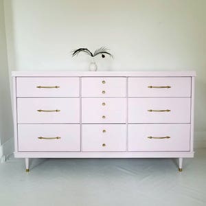 May include: A light pink chest of drawers with eight drawers and gold hardware. The dresser has a simple design with rounded edges and sits on four legs.