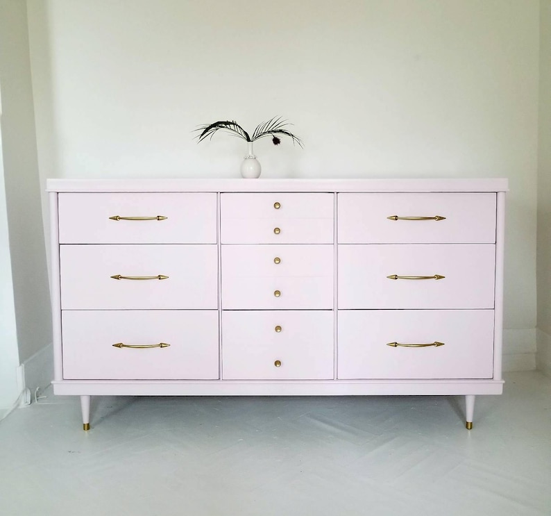 May include: A light pink chest of drawers with eight drawers and gold hardware. The dresser has a simple design with rounded edges and sits on four legs.