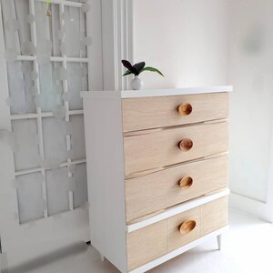 May include: White dresser with four drawers and a bottom shelf. The drawers have wood fronts and oval wooden knobs. The dresser has white legs.