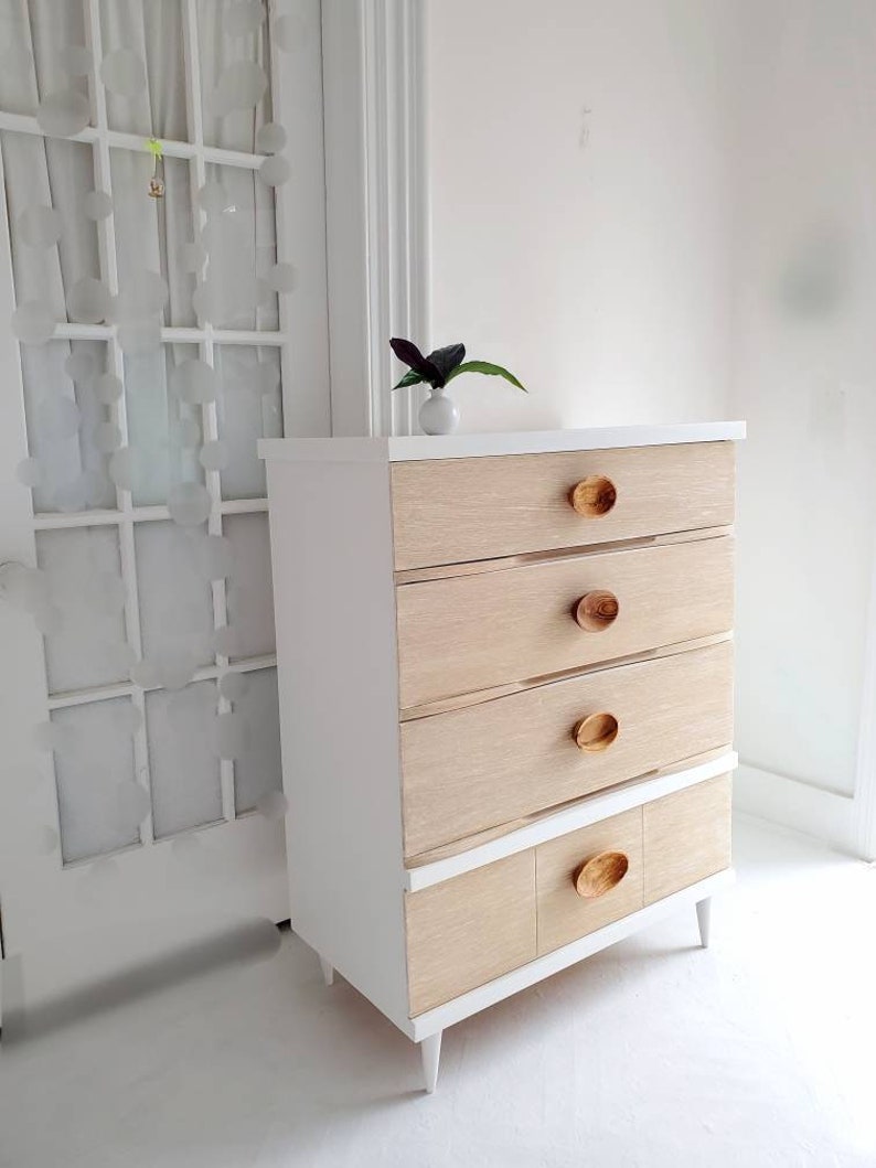 May include: White dresser with four drawers and a bottom shelf. The drawers have wood fronts and oval wooden knobs. The dresser has white legs.