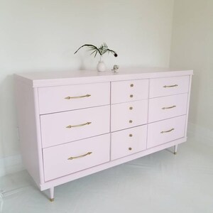 May include: A light pink dresser with eight drawers and gold hardware. The dresser has a modern design with sleek lines and a minimalist aesthetic.