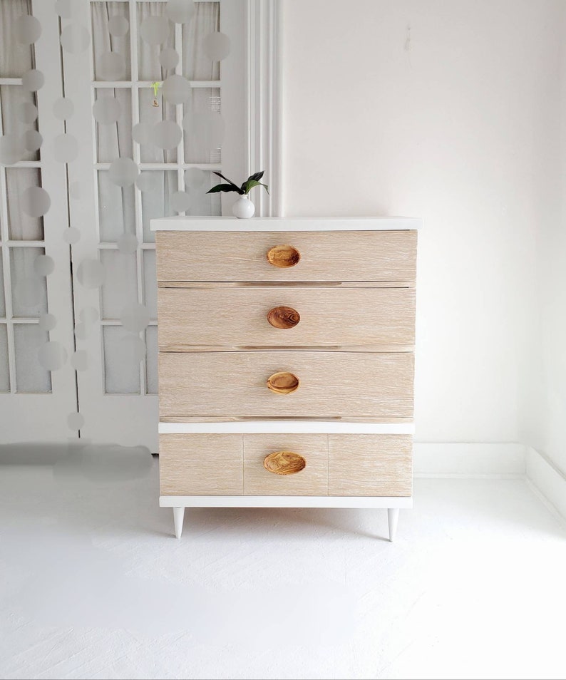 May include: A white dresser with five drawers and wooden knobs. The dresser has a light wood grain finish and sits on white legs.