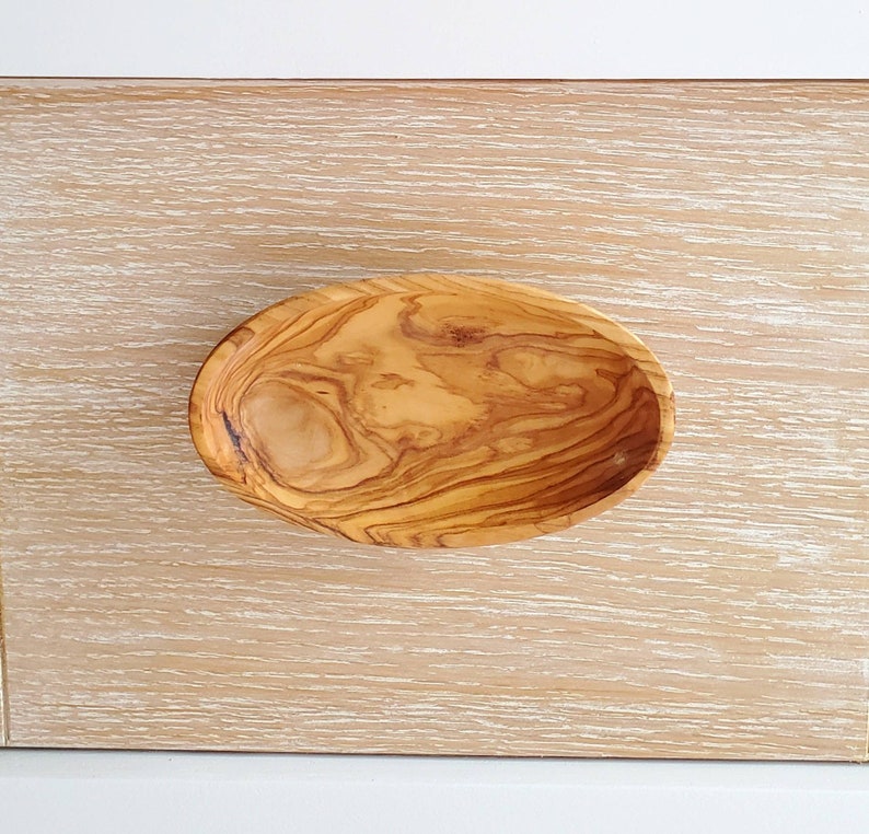 May include: A small, oval-shaped wooden bowl made from olive wood. The bowl has a natural wood grain pattern and is perfect for serving small snacks or dips.