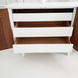 May include: White dresser with three drawers, the top drawer is open, revealing the wooden interior. The dresser has a wood grain finish on the sides and is sitting on a white floor.