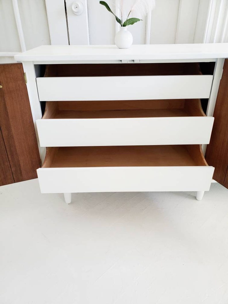 May include: White dresser with three drawers, the top drawer is open, revealing the wooden interior. The dresser has a wood grain finish on the sides and is sitting on a white floor.