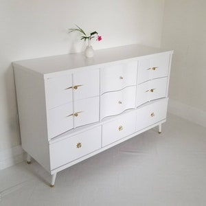 May include: White dresser with six drawers and gold hardware. The dresser has a curved front and sits on four legs.