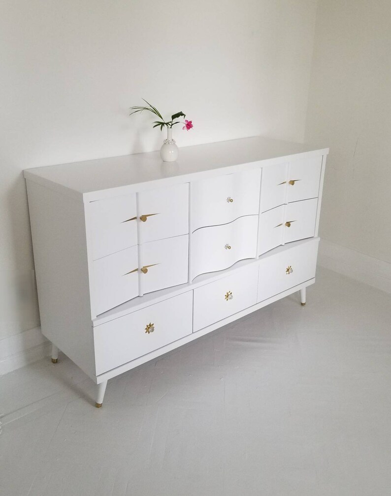 May include: White dresser with six drawers and gold hardware. The dresser has a curved front and sits on four legs.