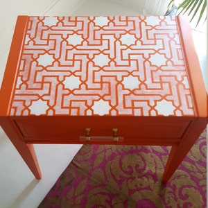 May include: Orange side table with a white geometric pattern on the top. The table has a single drawer with gold handles and sits on a pink and brown patterned rug.