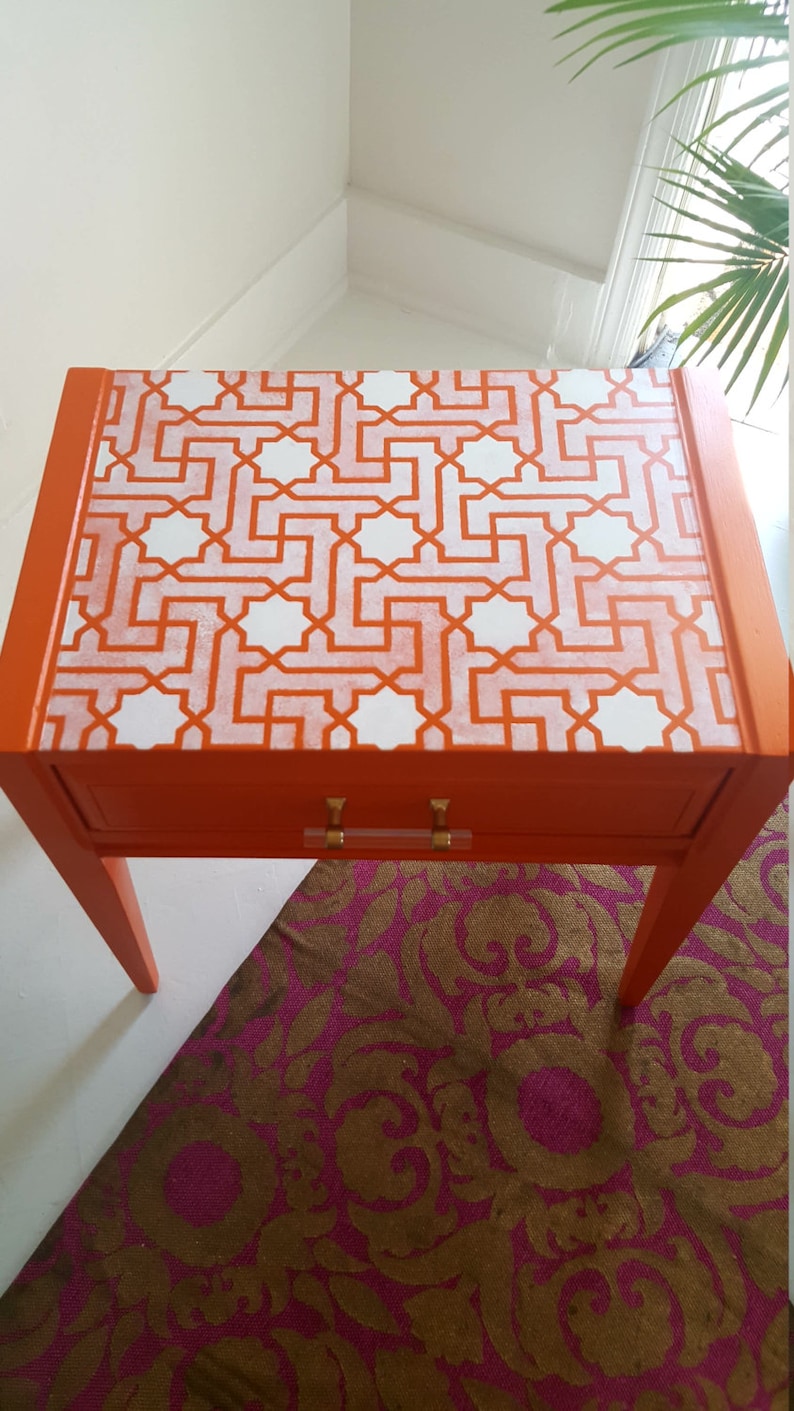 May include: Orange side table with a white geometric pattern on the top. The table has a single drawer with gold handles and sits on a pink and brown patterned rug.