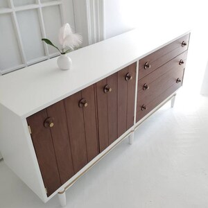May include: White mid-century modern dresser with brown wood cabinet doors and drawers. The dresser has gold trim and white legs.