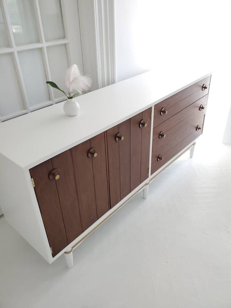 May include: White mid-century modern dresser with brown wood cabinet doors and drawers. The dresser has gold trim and white legs.