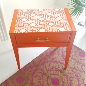 May include: Orange end table with a single drawer and a geometric patterned top. The table has gold-colored hardware and sits on a pink and gold patterned rug.