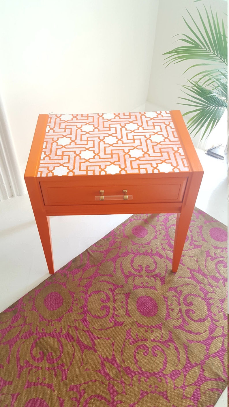 May include: Orange end table with a single drawer and a geometric patterned top. The table has gold-colored hardware and sits on a pink and gold patterned rug.